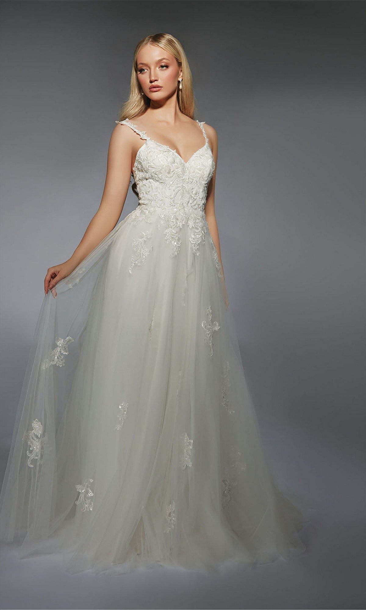A woman models the Alyce 7124 long white dress, an A-line wedding gown with lace details and lace appliques, standing against a gray background.